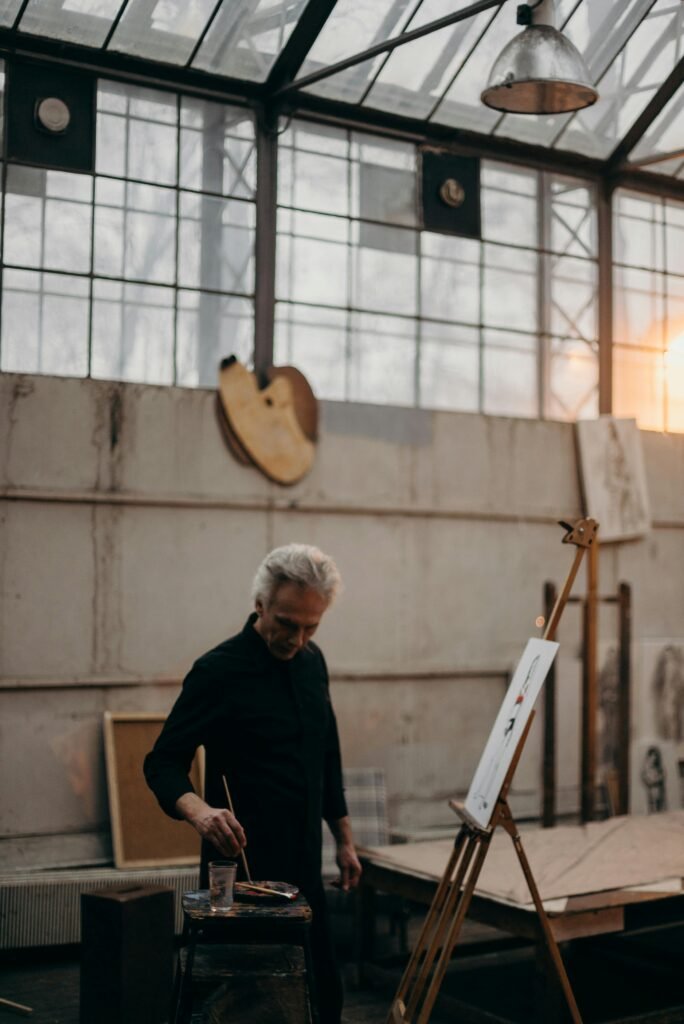 A senior male artist focused on painting in a rustic, sunlit studio with an easel.